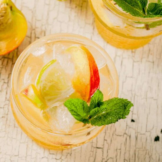 banner image of a glass of ice sangria made with roxyann winery rose. there is a peach and a sprig of mint on top of the glass. banner reads in permanent marker font "peachy sangria summer cocktail"