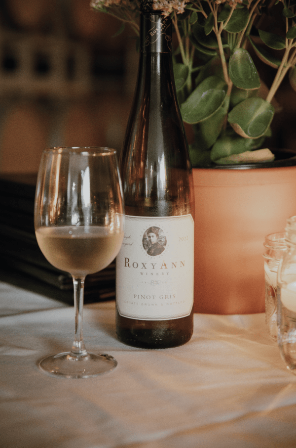 An image featuring a classic Oregon Winery bottle of Pinot Gris sitting next to a full glass of white wine. The label on the bottle reads "RoxyAnn Winery Pinot Gris."